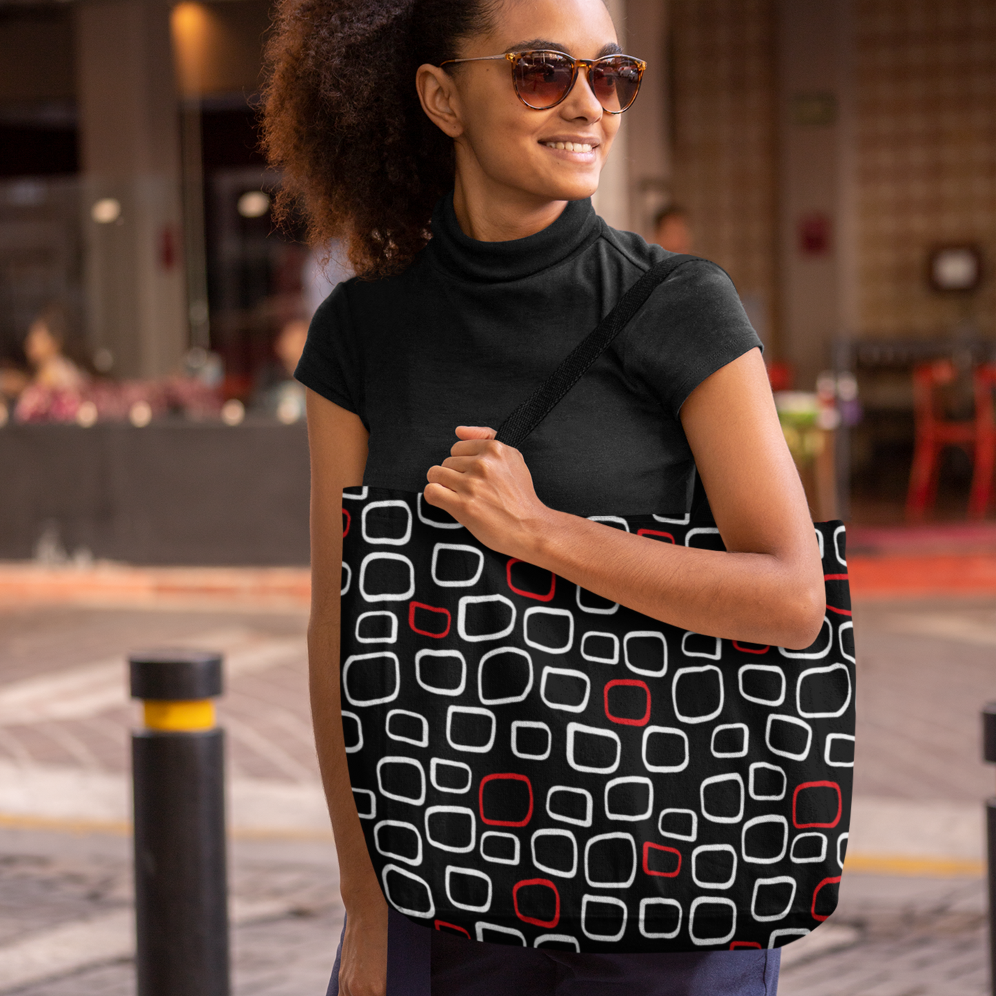Woman wearing sunglasses and smiling while holding a tote bag with a red, black, and white geometric pattern. She's outdoors with a relaxed vibe.