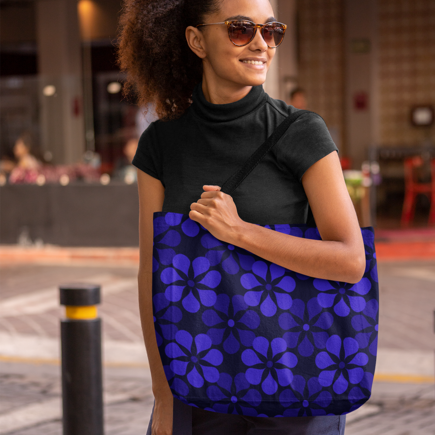 Smiling woman with curly hair and sunglasses holds a blue floral tote bag. She's wearing a black turtleneck, standing in a lively outdoor setting.