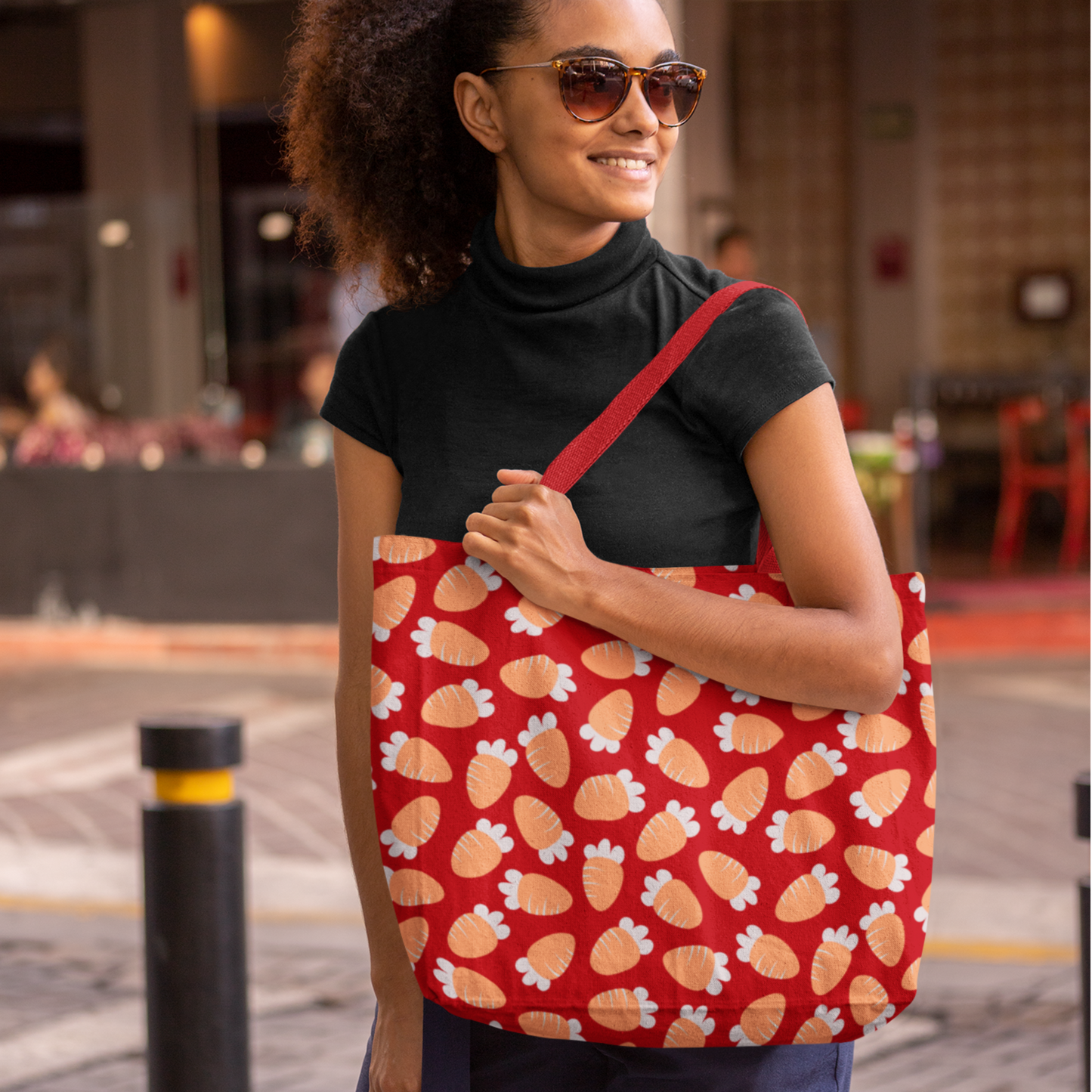 A woman in a black turtleneck and sunglasses smiles while holding a red tote bag with a carrot pattern. The background is an outdoor urban setting.