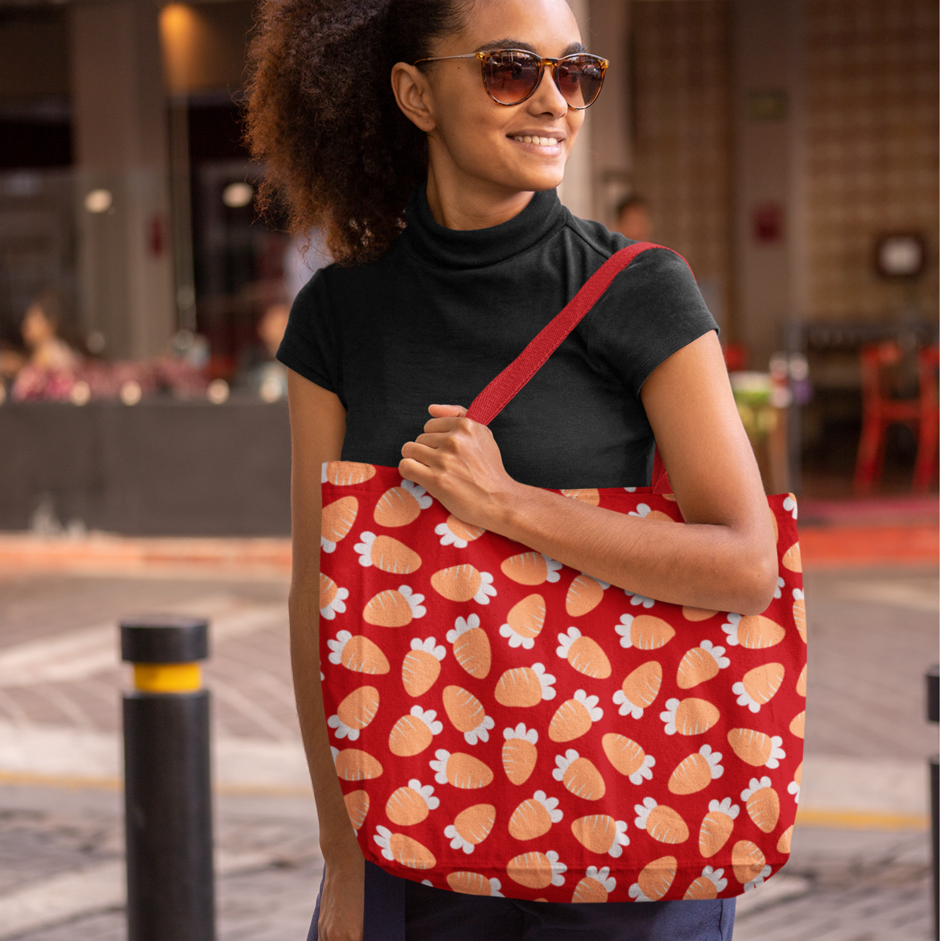 A woman in a black turtleneck and sunglasses smiles while holding a red tote bag with a carrot pattern. The background is an outdoor urban setting.
