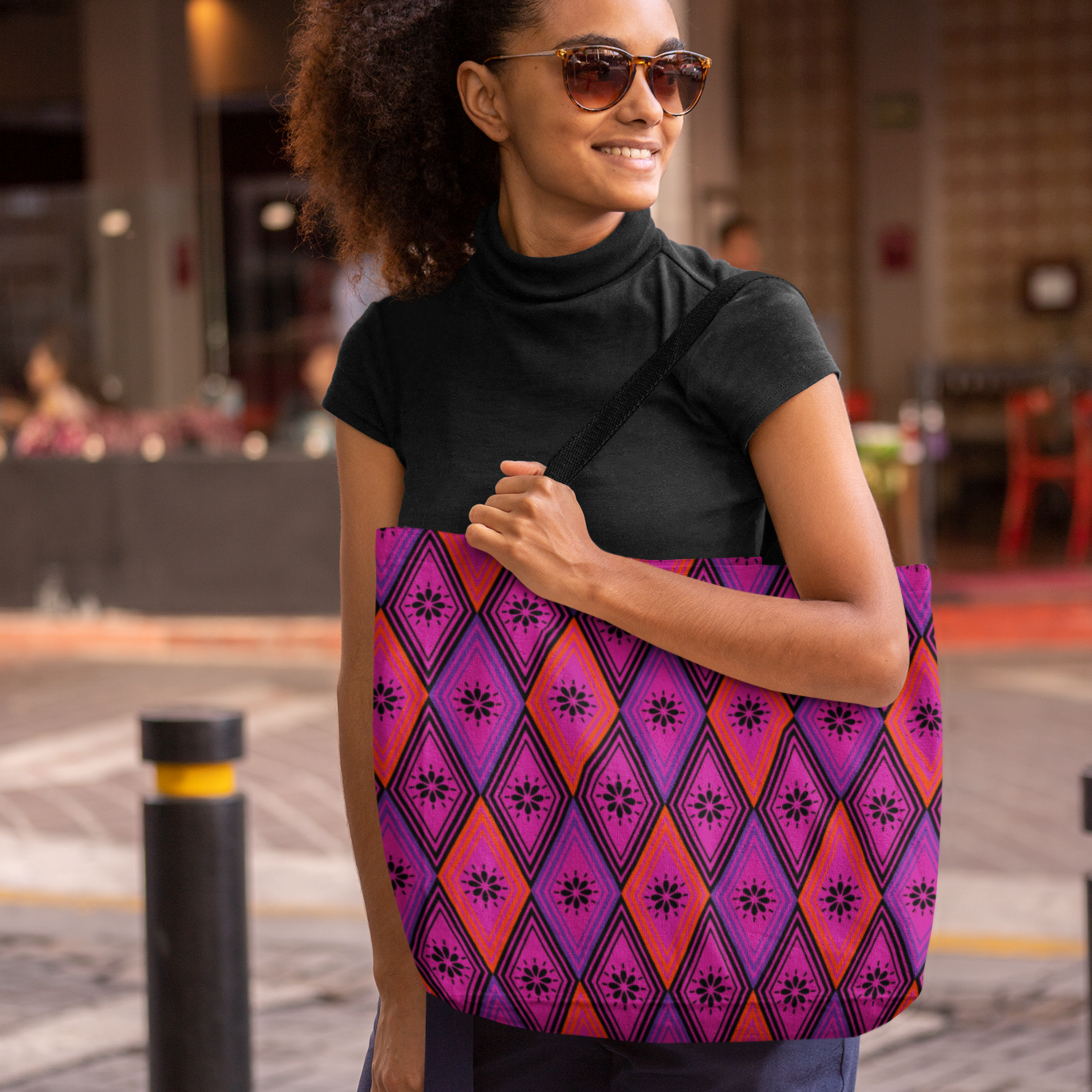  A woman in sunglasses smiles while carrying a vibrant tote bag featuring purple and pink diamond patterns. She's outdoors, creating a cheerful tone.